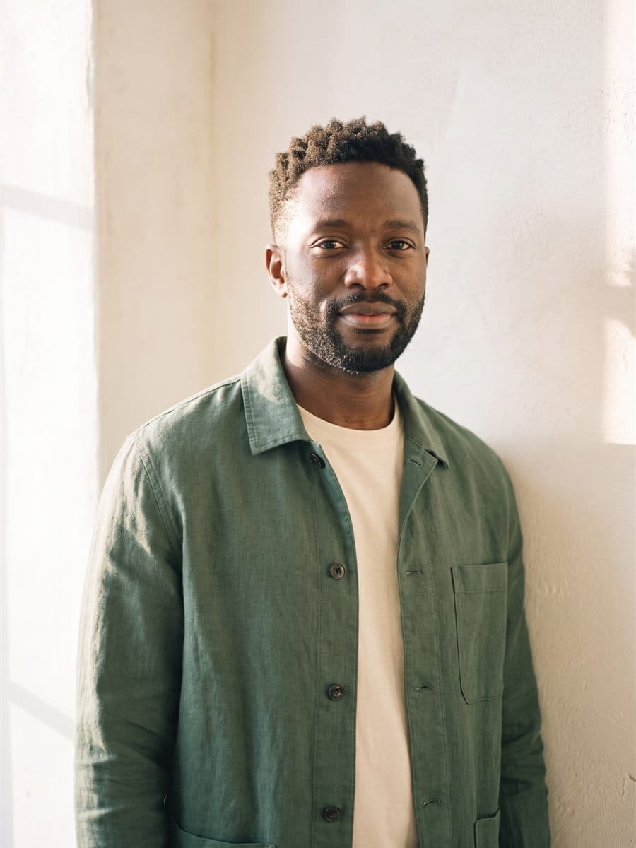 Devon Okafor in a deep sage chore jacket over a cream tee against a warm plaster wall, calm and settled