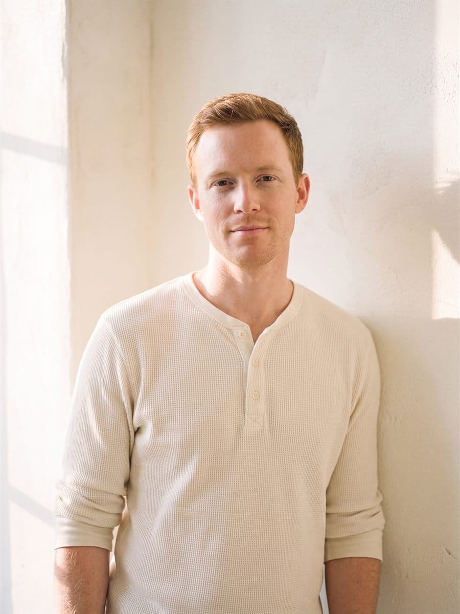 Eamon Walsh in a cream waffle-knit henley with pushed-up sleeves against a warm plaster wall, an easy near-smile