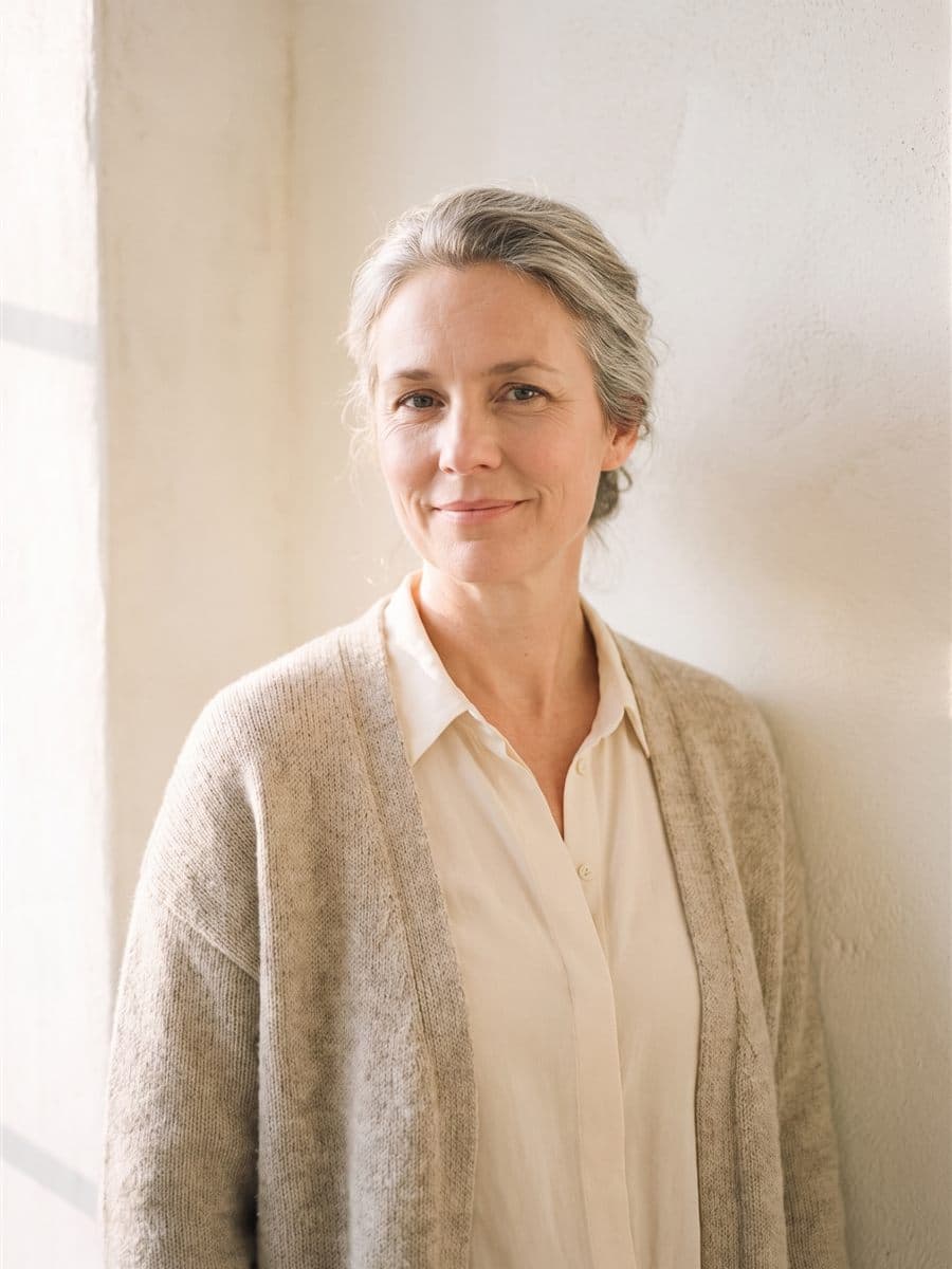 Ruth Neufeld in an oatmeal knit cardigan, silver-streaked hair in a loose bun, warm plaster wall and morning light