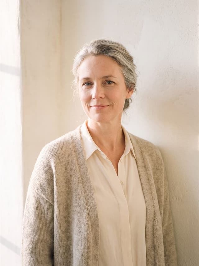 Ruth Neufeld in an oatmeal knit cardigan, silver-streaked hair in a loose bun, warm plaster wall and morning light