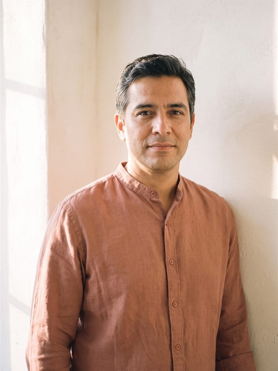 Tomás Vela in a muted terracotta band-collar shirt against a warm plaster wall, dark hair touched with grey